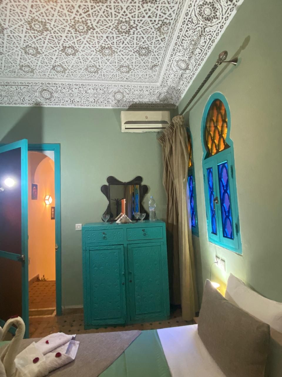 Una habitación con un armario verde azulado, una cama, un techo decorativo y una ventana con vidrio azulado.