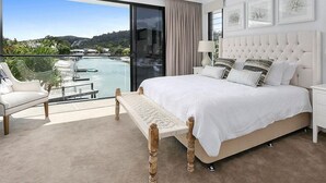 Un dormitorio moderno con una cama grande, ventilador de techo, vista a una piscina y montañas, y un balcón con una silla.