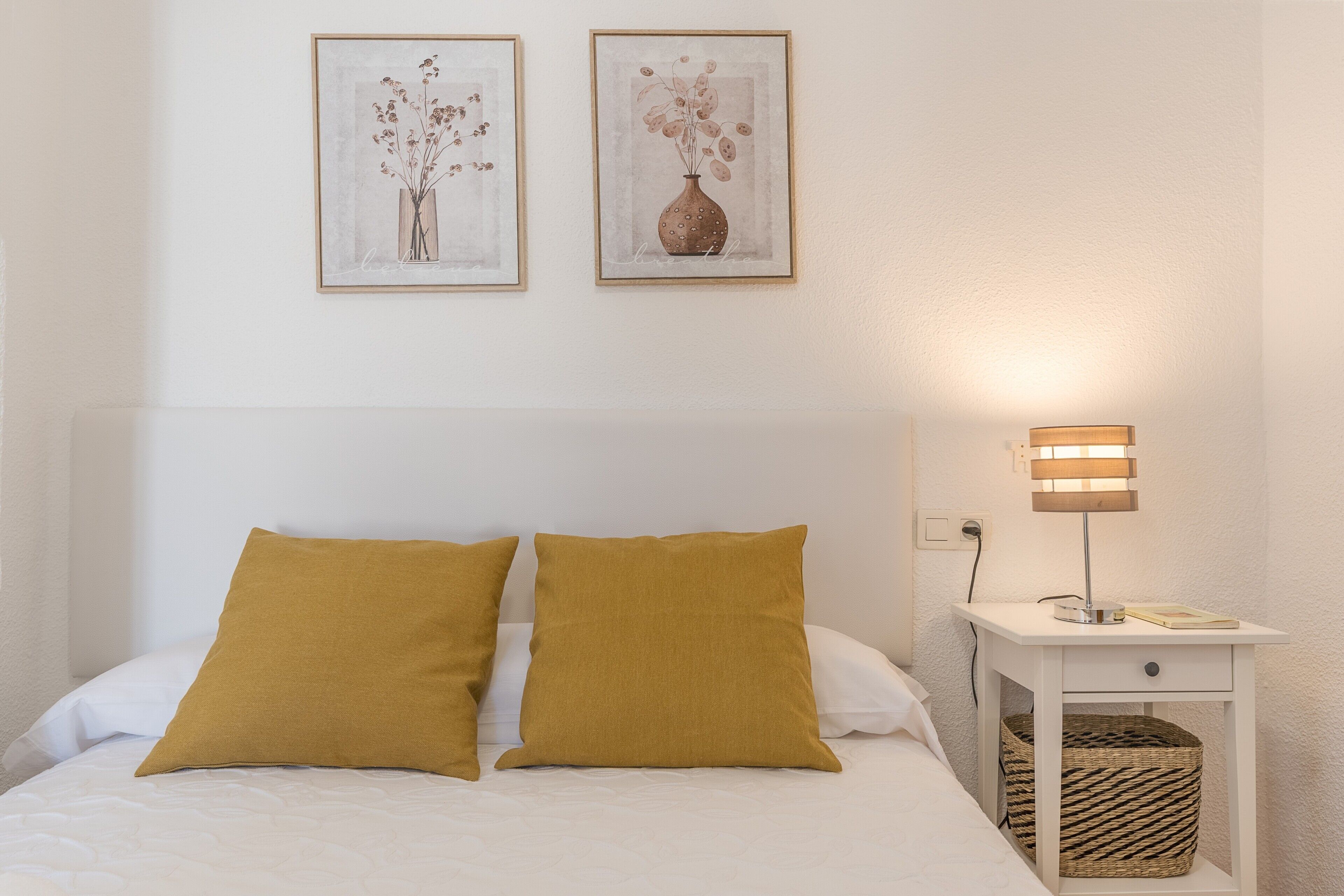 Een netjes opgemaakt bed met twee kussens, een nachtkastje met een lamp, en twee ingelijste foto’s aan de muur.
