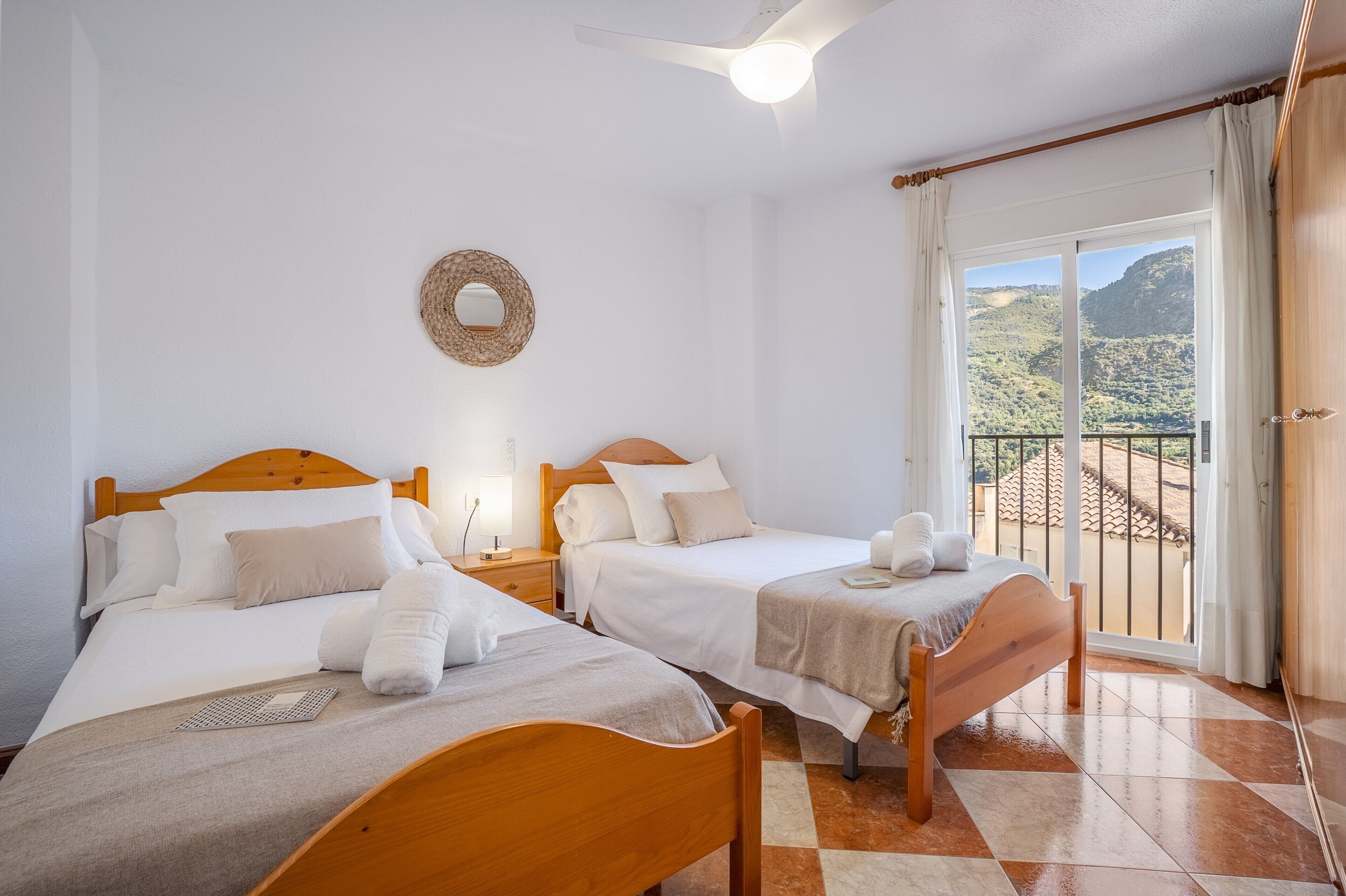 Un dormitorio con dos camas, un ventilador de techo, vistas a las montañas desde un balcón y un adorno de pared tejido.