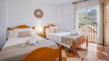 Un dormitorio con dos camas, un ventilador de techo, vistas a las montañas desde un balcón y un adorno de pared tejido.