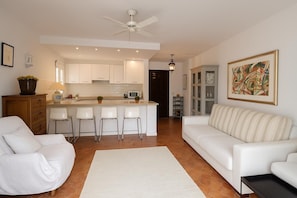 A modern kitchen with white cabinets, a central island with bar stools, a ceiling fan, and a dining area with a sofa and chairs.