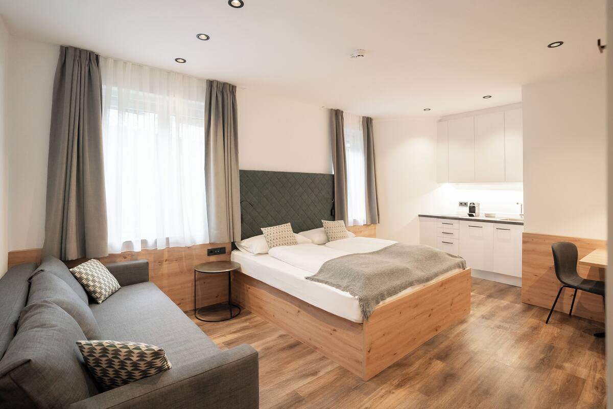 Une chambre à coucher moderne avec un lit en bois, un canapé gris, une petite table ronde et un coin cuisine.