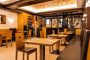 Restaurante com mesas e cadeiras de madeira, área de bar e um espaço com bancos estofados.