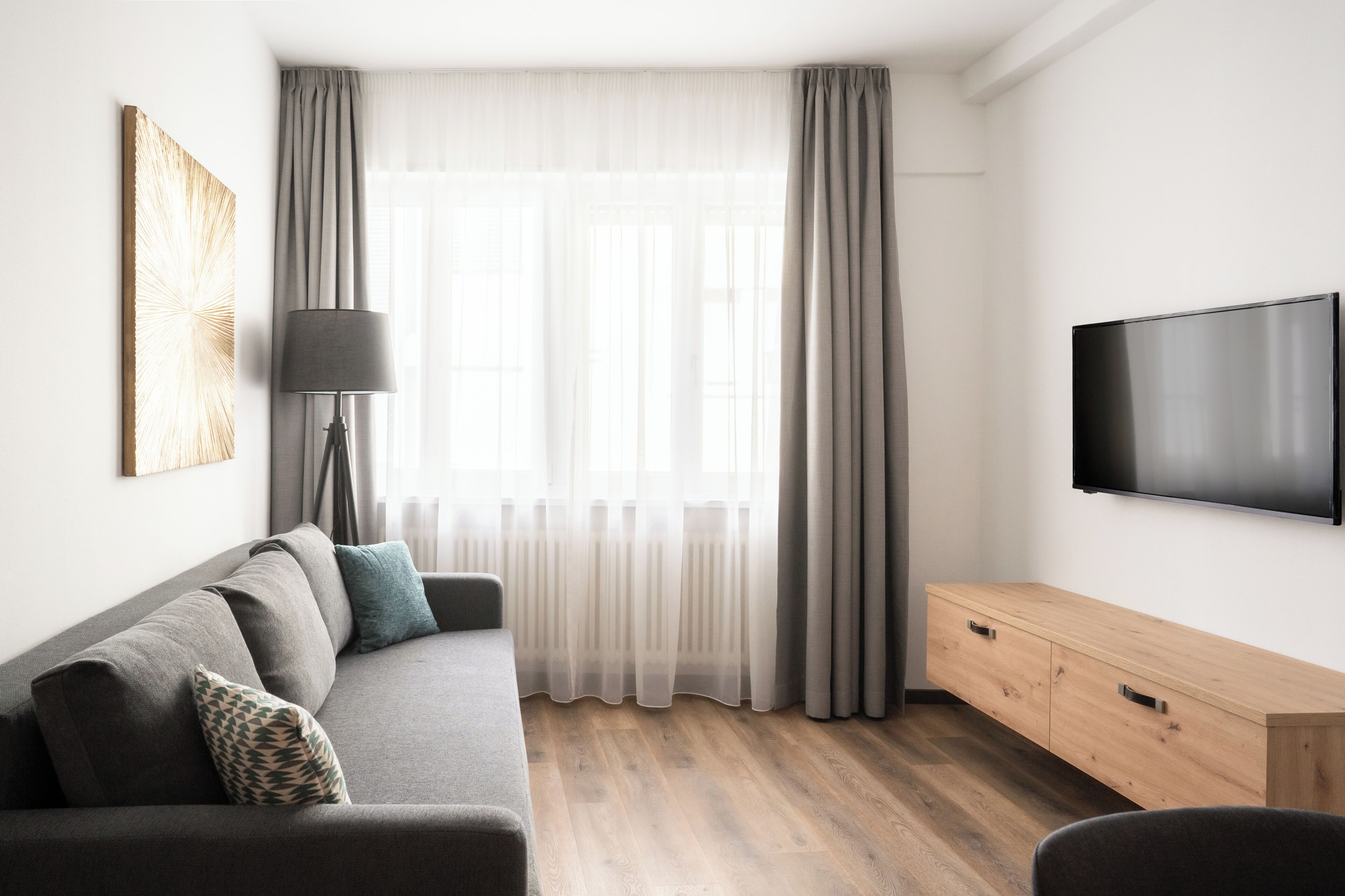 A modern living room with a grey sofa, a wooden TV stand, and a large window with sheer curtains.