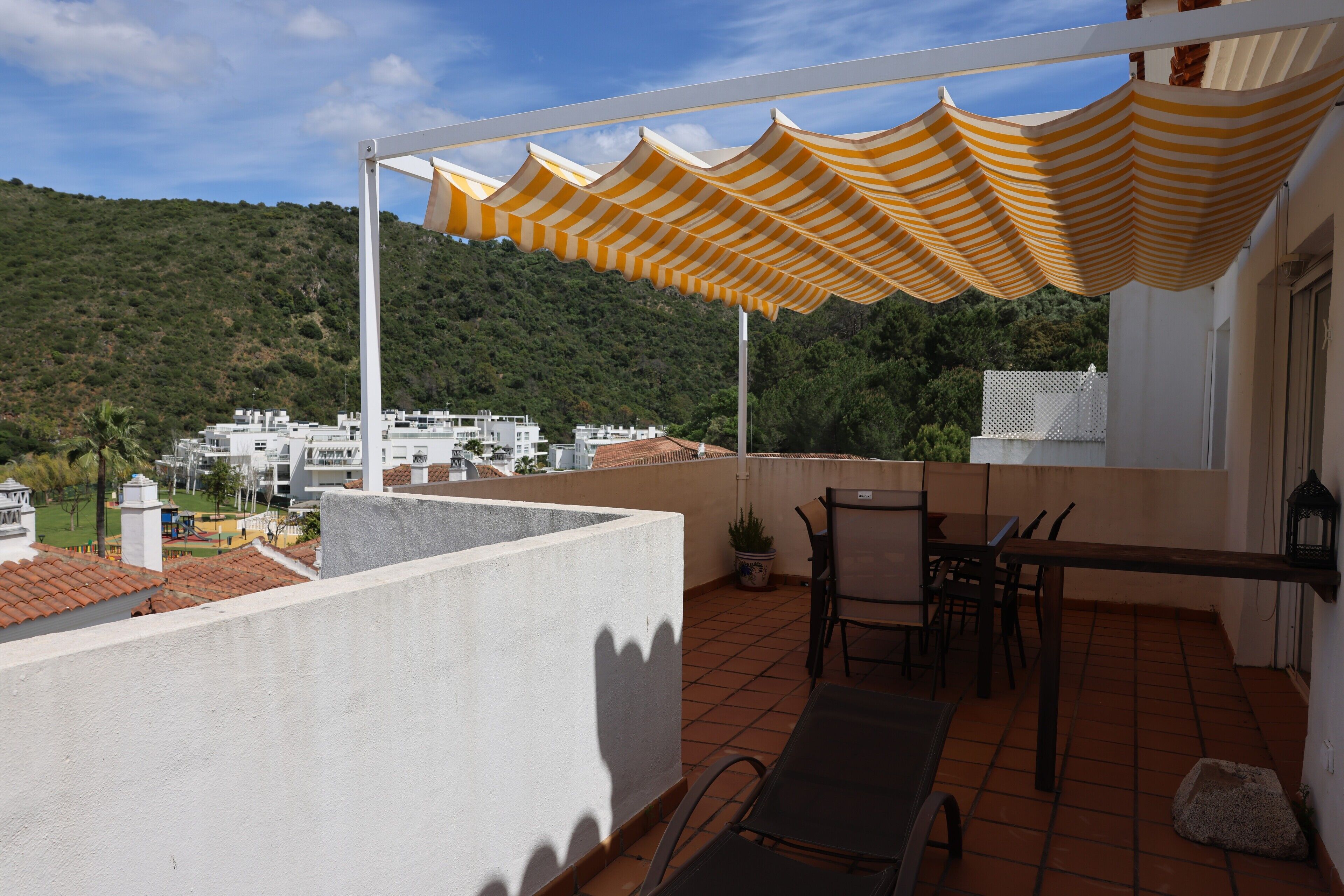 A terrace with a table and chairs, a potted plant, and a view of a hillside with buildings.