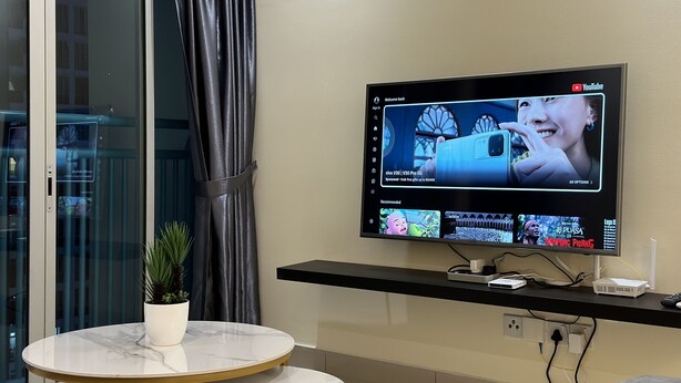 En moderne stue med et fladskærms-tv, der viser en YouTube-kanal, et marmorbord med en potteplante og et sort konsolbord med elektroniske enheder.