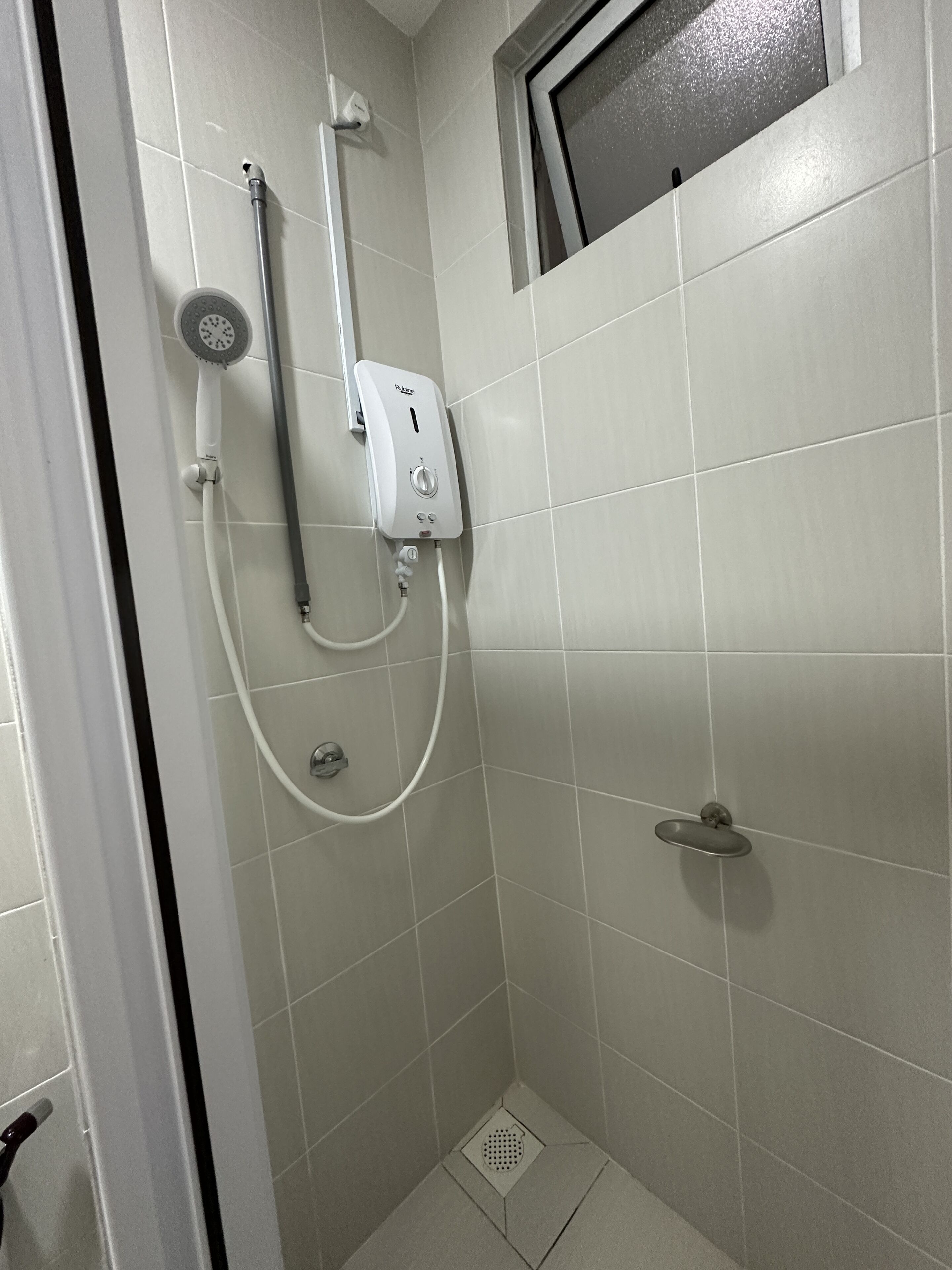 A small, tiled shower area with a handheld showerhead, a wall-mounted control panel, and a handheld shower hose.