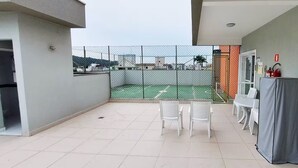 A balcony with a view of a tennis court, white chairs, and a covered table. - Complete apartment in Ubatuba - Itaguá with air conditioning + Wifi (Itaguá Ubatuba)
