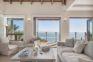 A modern living room with a large window overlooking the sea, a sofa, a coffee table with books, and a view of the ocean. - Luxe Oceanfront Villa | The Chosen Collection (Goleta)