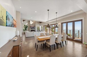 Un vaste espace de salle à manger avec une table et des chaises en bois, une cuisine à l’arrière-plan et une vue sur la mer grâce à de grandes portes coulissantes.