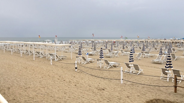A beach with rows of white lounge chairs and striped umbrellas, a sandy shore, and a calm sea in the background.