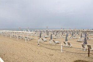 A beach with rows of white lounge chairs and striped umbrellas, a sandy shore, and a calm sea in the background.