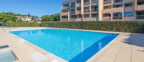 A rectangular swimming pool with a tiled deck, surrounded by a low hedge, and adjacent to a multi-story building with balconies.