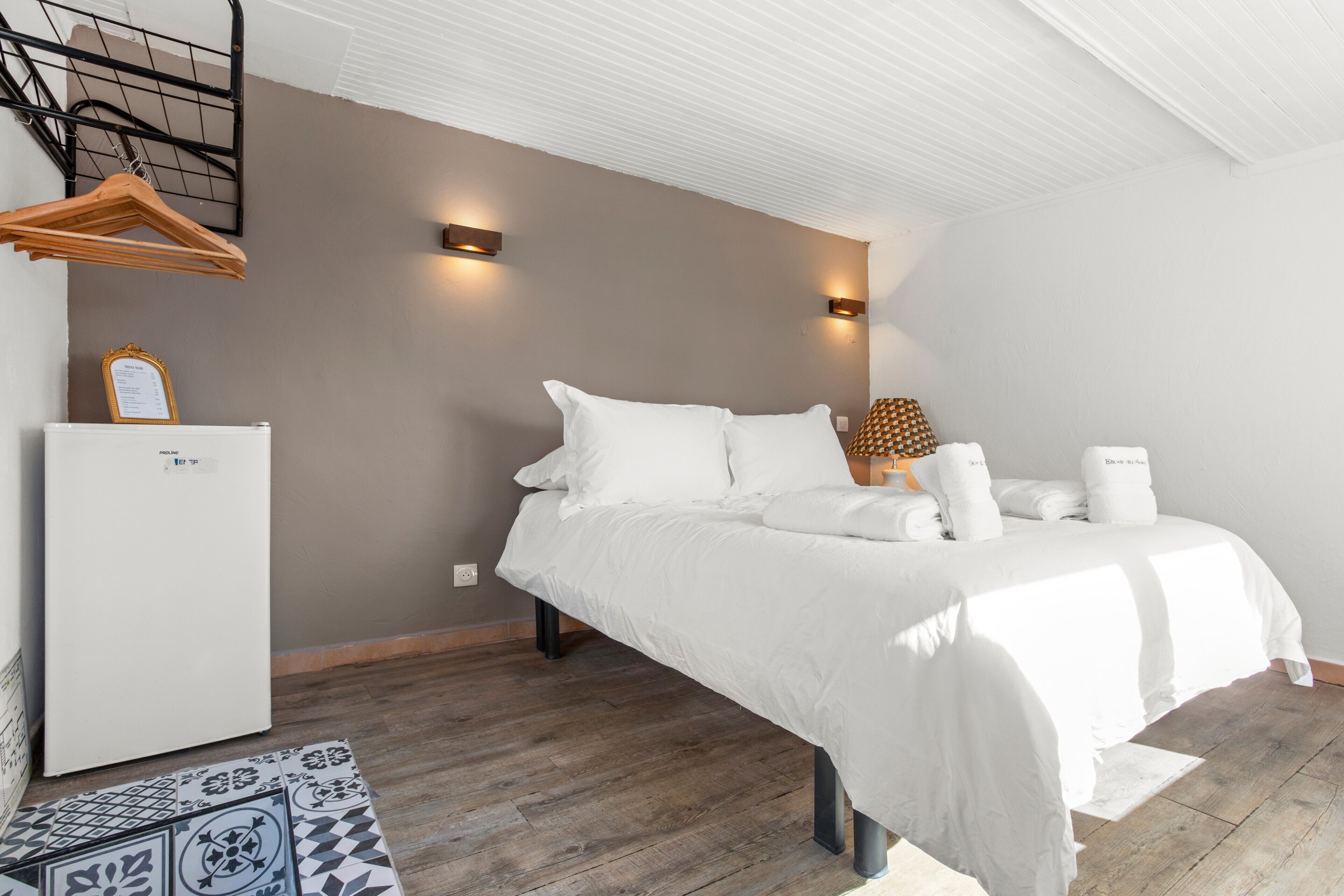 A bedroom with a bed, bedside lamps, a hanging rack, and a refrigerator.