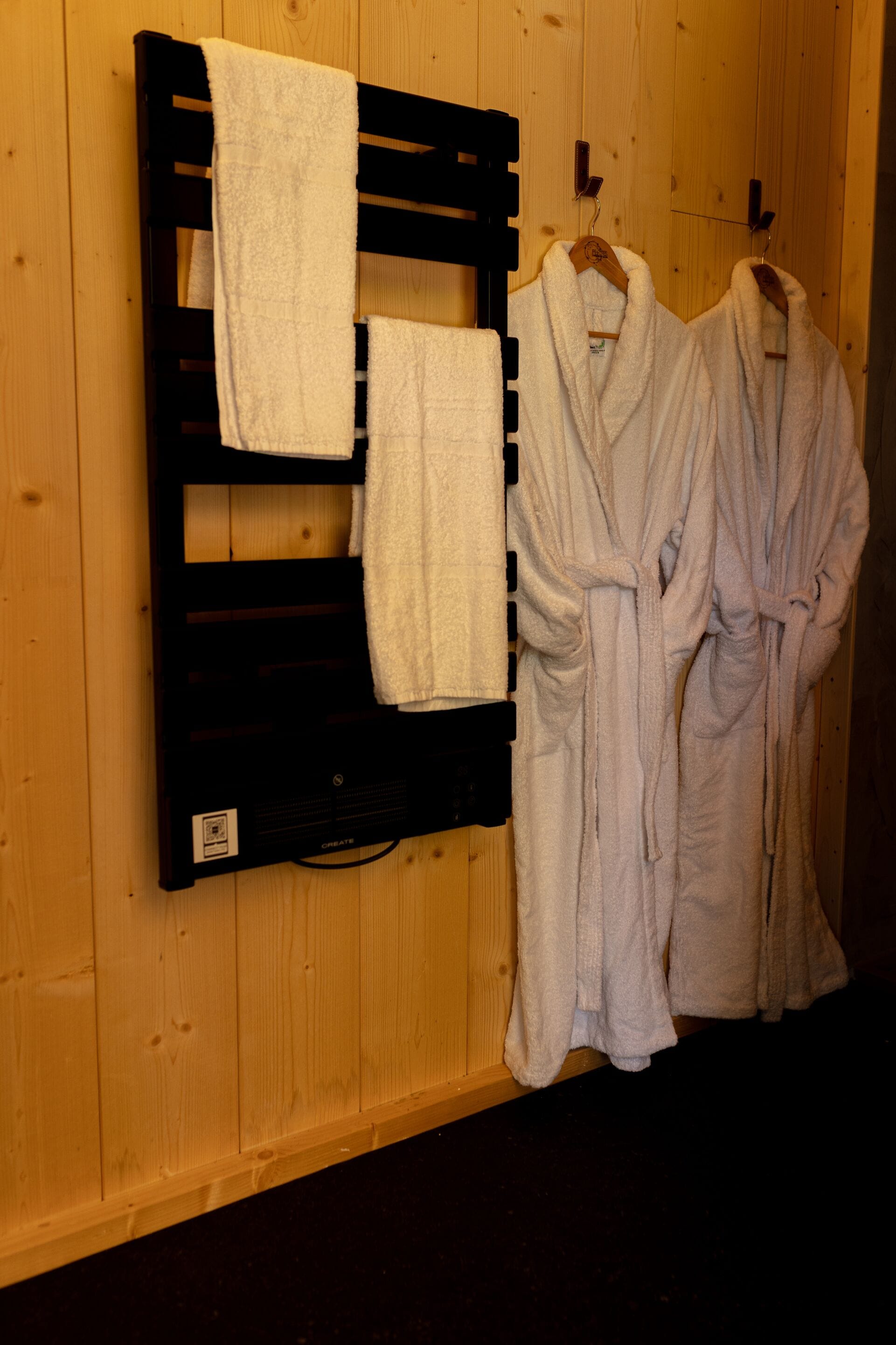 A wooden wall with a towel rack holding two white bathrobes and a towel.