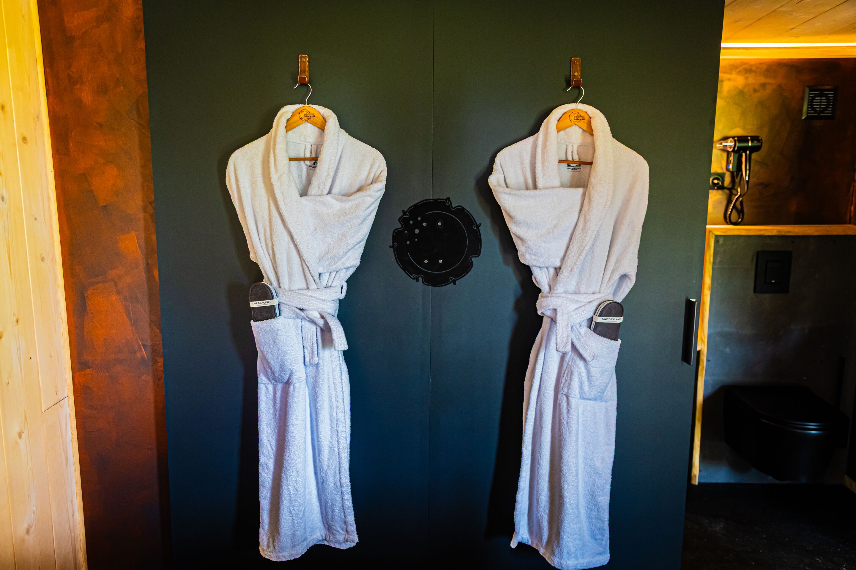 Two white robes hanging on a dark door with a towel rack, a black circular object, and a shower area visible to the right.