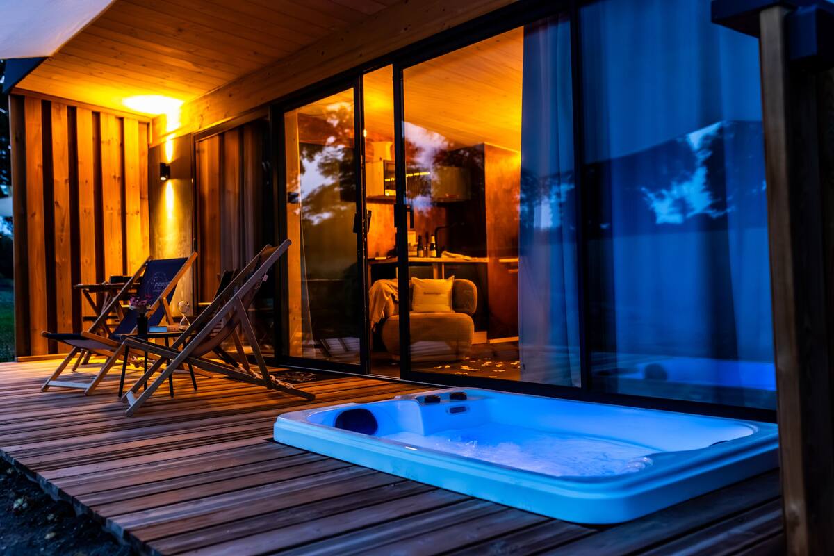 A wooden deck with a hot tub, lounge chairs, and a glass-enclosed living area with a view of the outdoors.