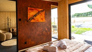 A modern room with a wooden ceiling, a large abstract painting on the wall, a bed with rolled towels, and a view of an outdoor area through a glass door. - Pépites Lodges et Spas (Prémilhat)