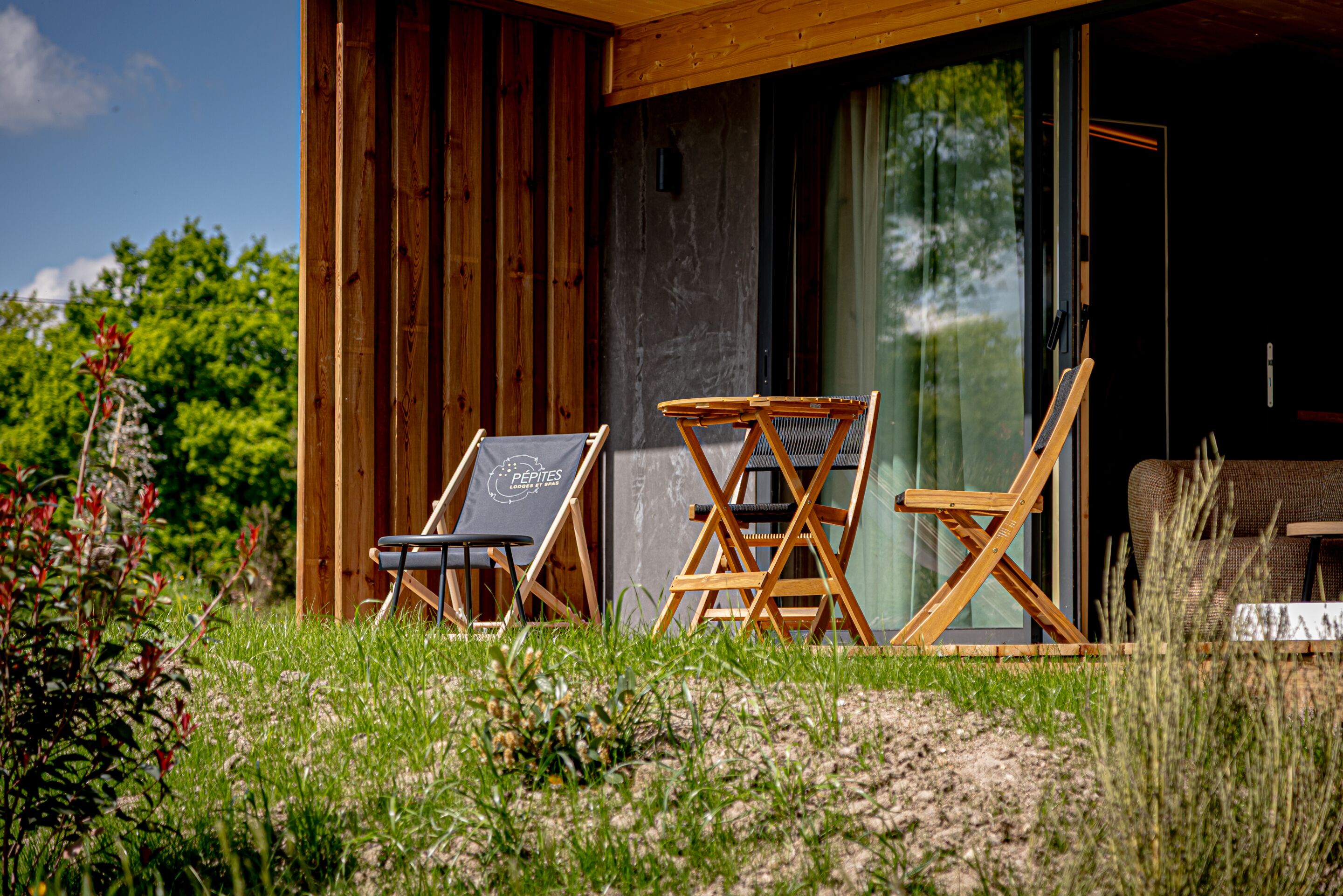 A wooden cabin with a glass door, two folding chairs, and a small table outside.
