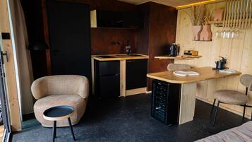 Cocina compacta con refrigerador integrado, un pequeño comedor con mesa de madera y dos sillas, y una zona de estar con silla de mimbre y un sofá.