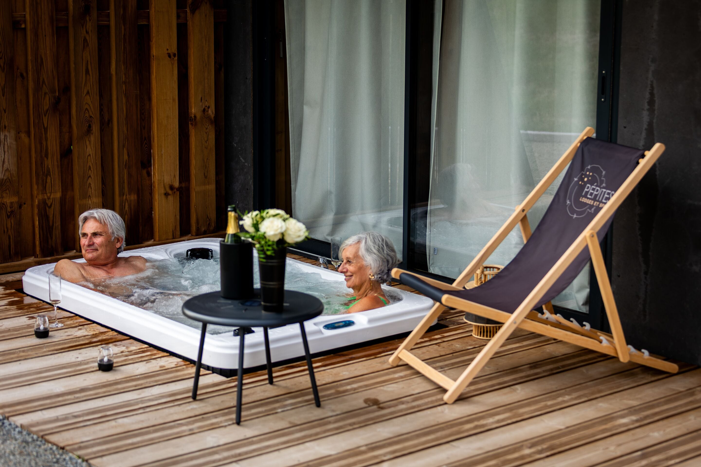 A hot tub with two people, a wooden deck, and a lounge chair.