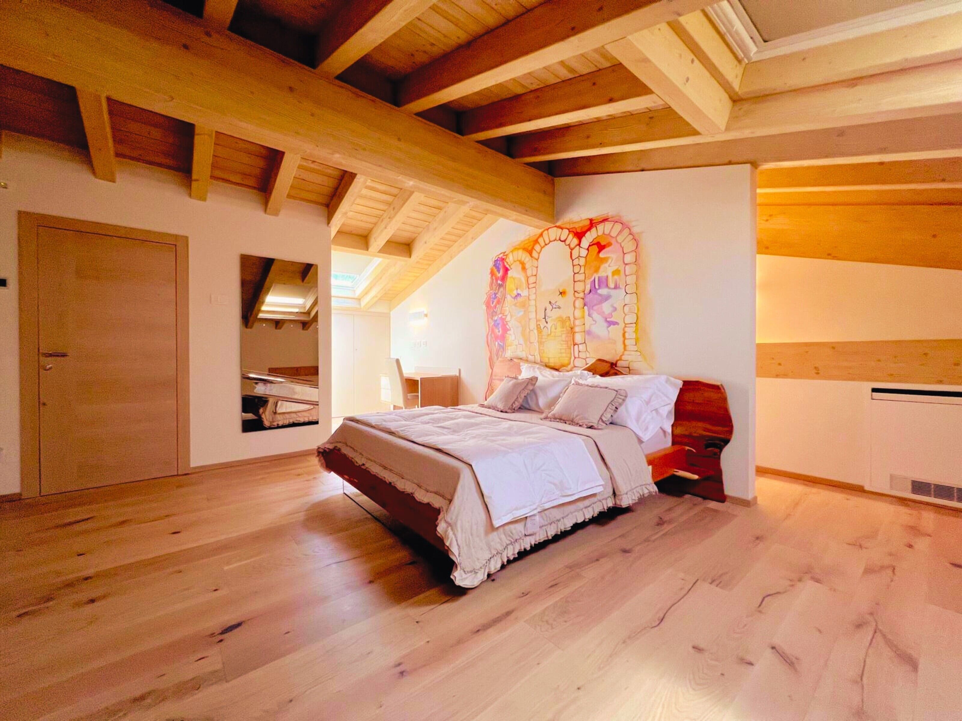Un dormitorio con techo de madera, una cama con ropa de cama blanca, suelo de madera y una pared decorada con una obra de arte colorida.