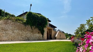 Un edificio de piedra con una escalera que conduce a un porche cubierto, rodeado de césped cuidado y plantas con flores.