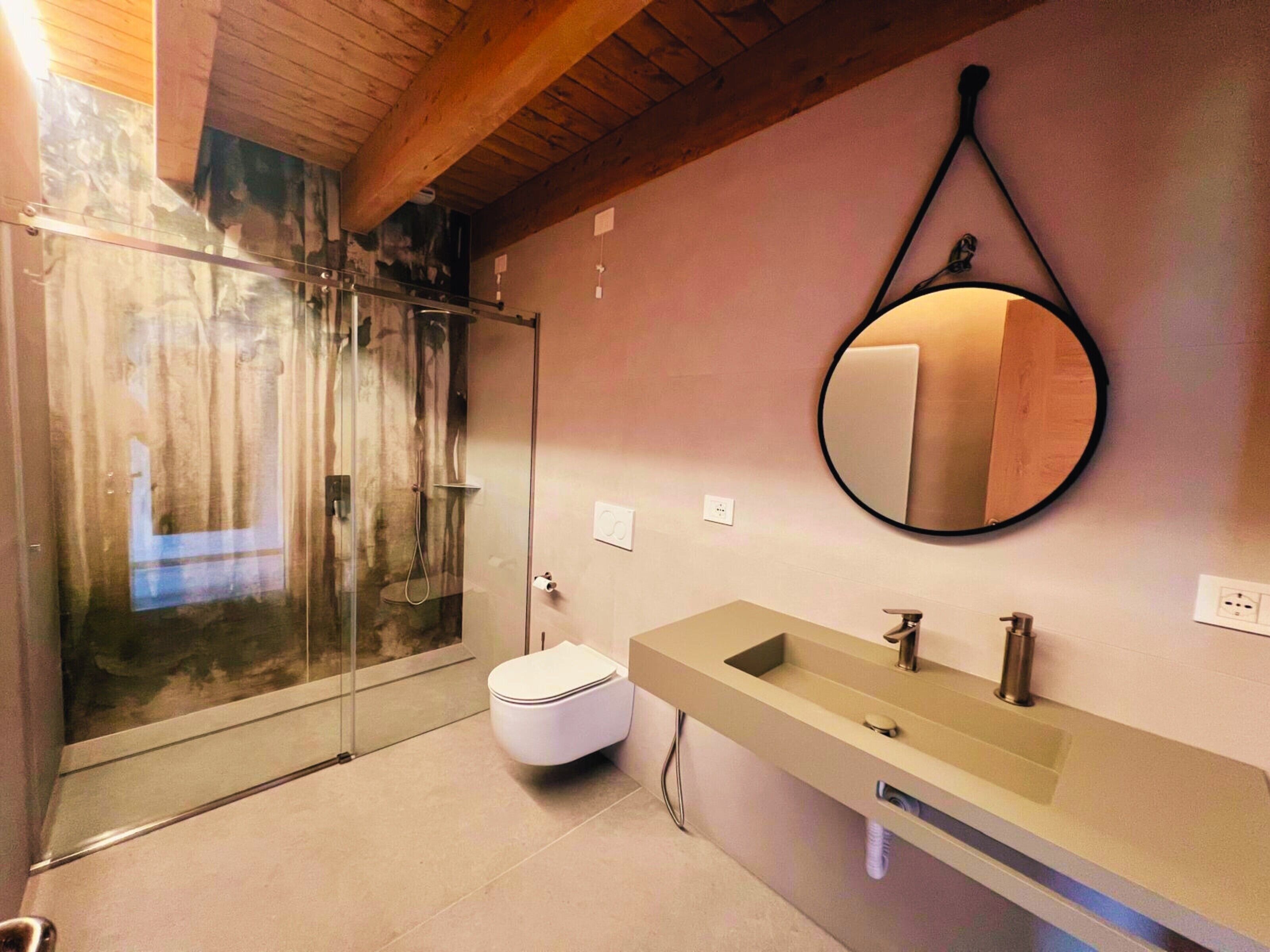 A modern bathroom with a glass shower enclosure, a white toilet, a beige sink with a mirror, and a wooden ceiling.