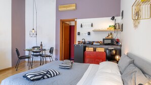 A bedroom with a bed, pillows, a bedside table, a desk with chairs, a kitchen area with a microwave, and a red cabinet. - Studio 'Il Tuo Rifugio' with Balcony, Wi-Fi and Air Conditioning (Modica)