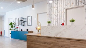A reception area with a wooden reception desk, a blue counter, and a wall with a tree pattern. There are framed pictures on the wall, a hanging pendant light, and potted plants on the counter.