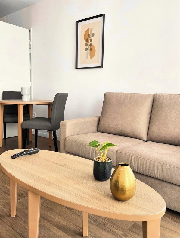 A modern living room with a beige sofa, a wooden coffee table, a remote control, a potted plant, and a decorative vase. - Zenao Appart'hôtels Wintzenheim (Wintzenheim)
