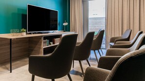 A modern room with a large flat-screen TV, a wooden desk, a bookshelf, and several upholstered chairs.