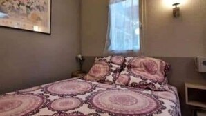 A bedroom with a bed, a window with a curtain, a wall-mounted light, and a painting on the wall. - Apartment 'Proche Mer' with Shared Pool, Wi-Fi and Air Conditioning (Argelès-sur-Mer)