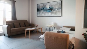 A modern living room with a sofa, a small table, a baby chair, and a bed with a blanket. - Lovely 1BR in Old Town: City Center & 4min to Beach (Larnaca)