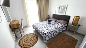 Quarto com cama, escrivaninha, cadeira, mesa de cabeceira, abajur, tapete e um quadro na parede.