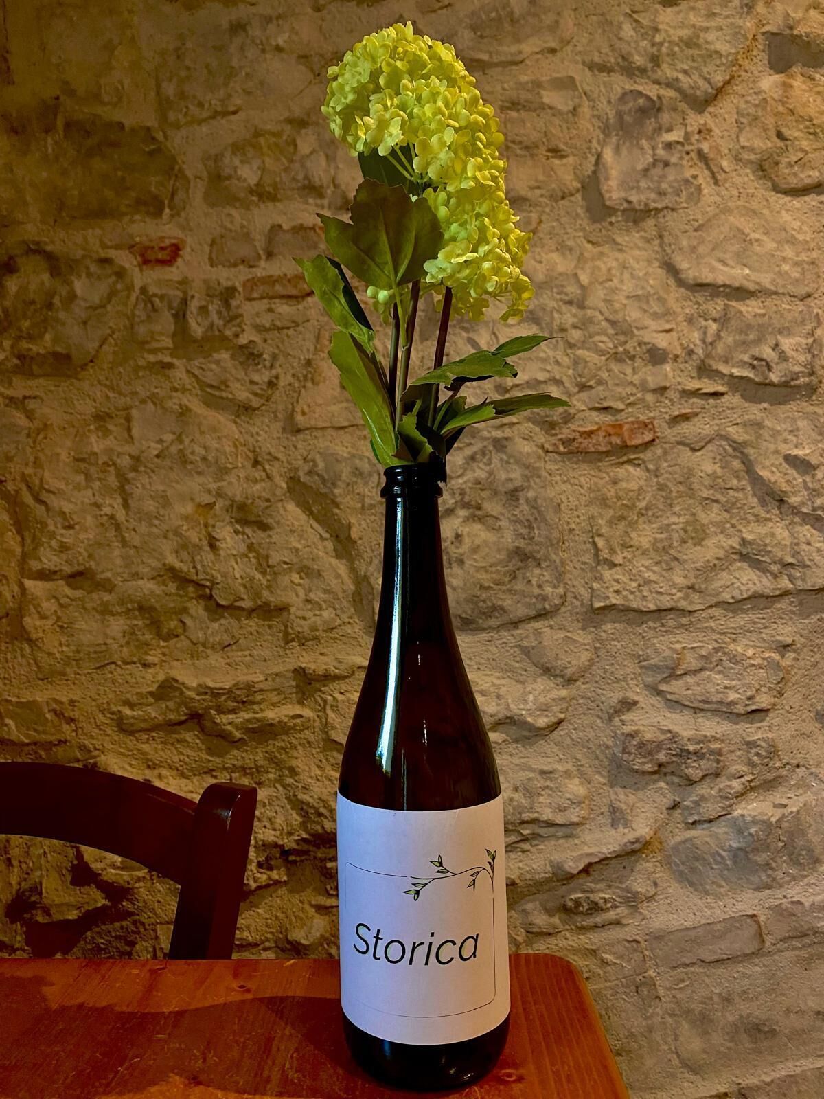 Una botella de vino con la etiqueta 'Storica' y un ramo de flores amarillas encima, sobre una mesa de madera frente a un fondo de pared de piedra.