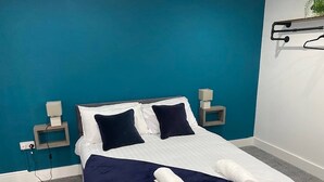 A neatly made bed with a dark blue blanket and pillows, a white headboard, and two bedside tables with lamps. - Apartment 07 St Andrews (Lytham St. Anne's)