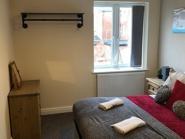 A bedroom with a bed, a window, a radiator, a small table, and a shelf. - 04 St Andrews - Sleeps 6 (Lytham St. Anne's)