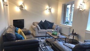 A living room with a sofa, ottoman, and a coffee table. There is a television on a stand and a window with a view of the outside. - 04 St Andrews - Sleeps 6 (Lytham St. Anne's)