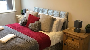 A neatly made bed with a red and grey blanket, multiple pillows, and a wooden nightstand with a lamp. - 04 St Andrews - Sleeps 6 (Lytham St. Anne's)