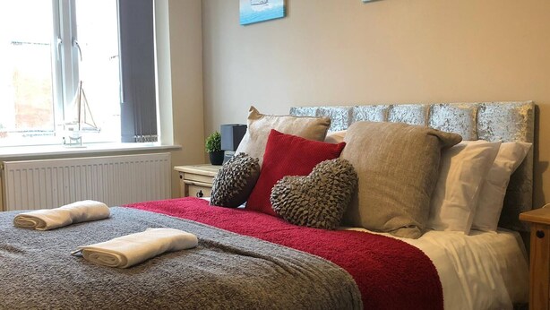 A neatly made bed with a red blanket and pillows, a small bedside table with a lamp, and a window with a view of a sailboat. - 04 St Andrews - Sleeps 6 (Lytham St. Anne's)