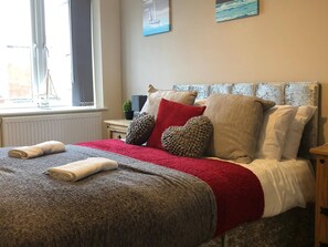 A neatly made bed with a red blanket and pillows, a small bedside table with a lamp, and a window with a view of a sailboat. - 04 St Andrews - Sleeps 6 (Lytham St. Anne's)