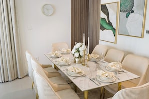 Un coin repas avec une longue table dressée pour six personnes, des chaises blanches et une horloge au mur.