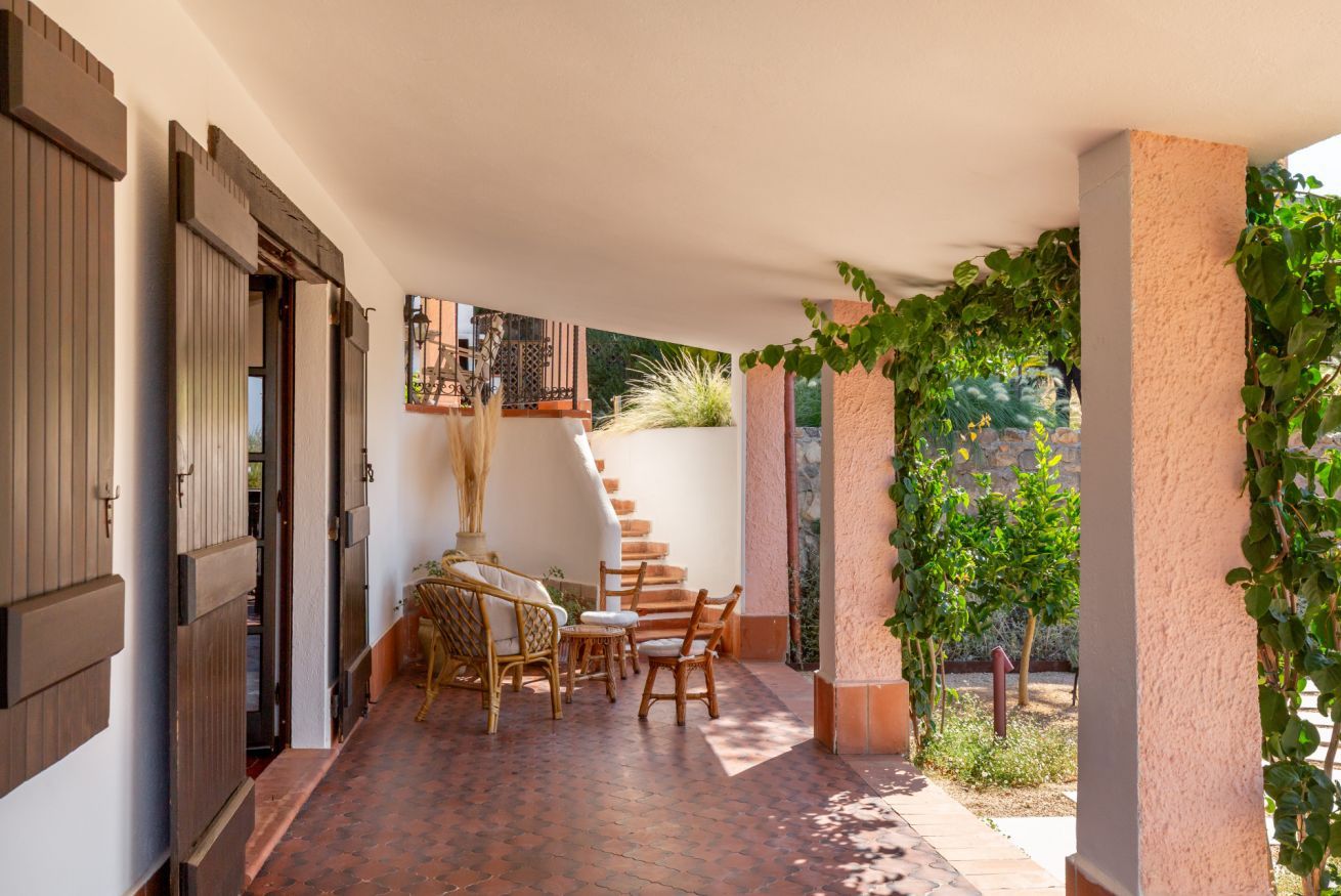 Un espacio al aire libre cubierto con piso de ladrillo, sillas de madera, una mesa y una escalera que conduce a un nivel superior.