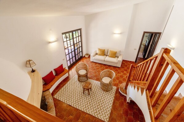 A spacious living area with a white sofa, two wicker chairs, a herringbone-patterned rug, and a wooden staircase.