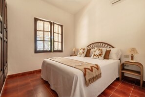 A bedroom with a bed, a window with a decorative pattern, a nightstand with a lamp, and a wall-mounted air conditioner.