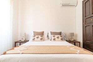 Un lit bien fait, recouvert d’une couvre-lit beige et blanche, avec deux oreillers assortis et une table de chevet en bois sur laquelle se trouve une lampe.