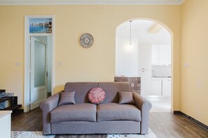 Sala de estar com sofá, um prato decorativo e vista para uma cozinha através de um arco.