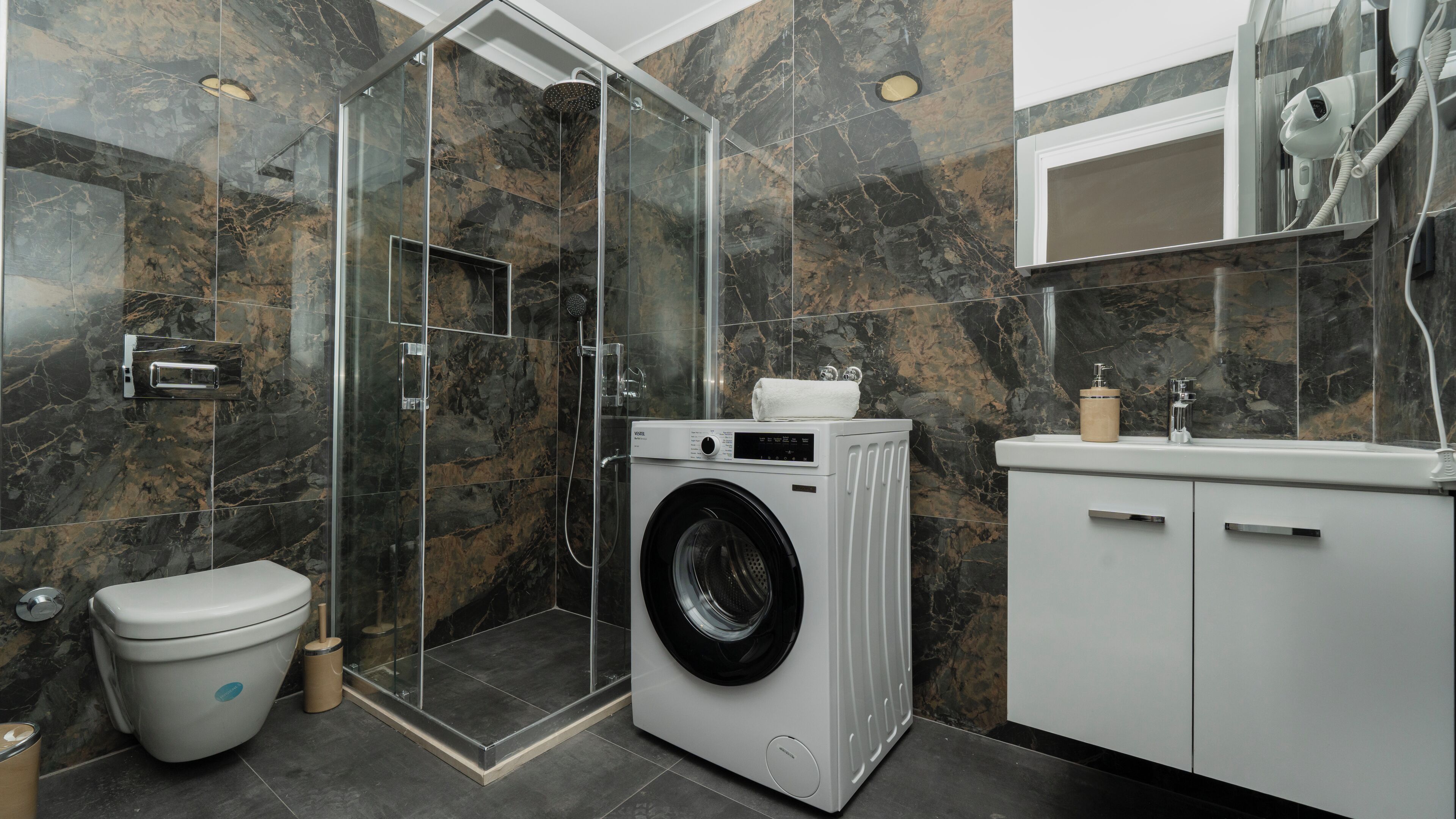 A modern bathroom with a glass-enclosed shower, a built-in washing machine, a toilet, and a vanity with a mirror.